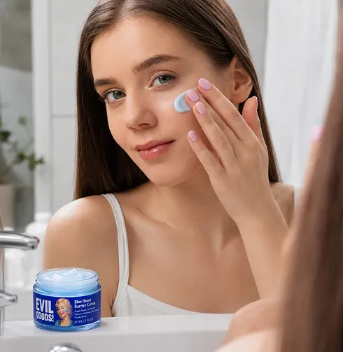 A woman applies blue face cream to her cheek while looking in a mirror, with the product jar nearby.