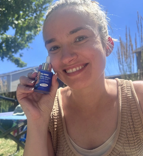 A smiling woman holds a small blue bottle of Blue Honey Peptide Serum next to her face outdoors.