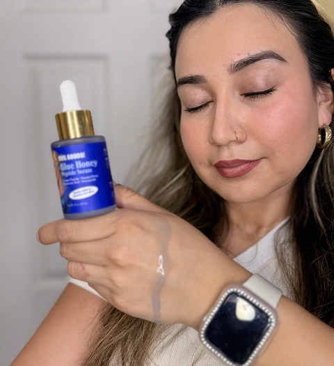 A woman with her eyes closed holds a blue bottle of serum with a swatch on her hand.