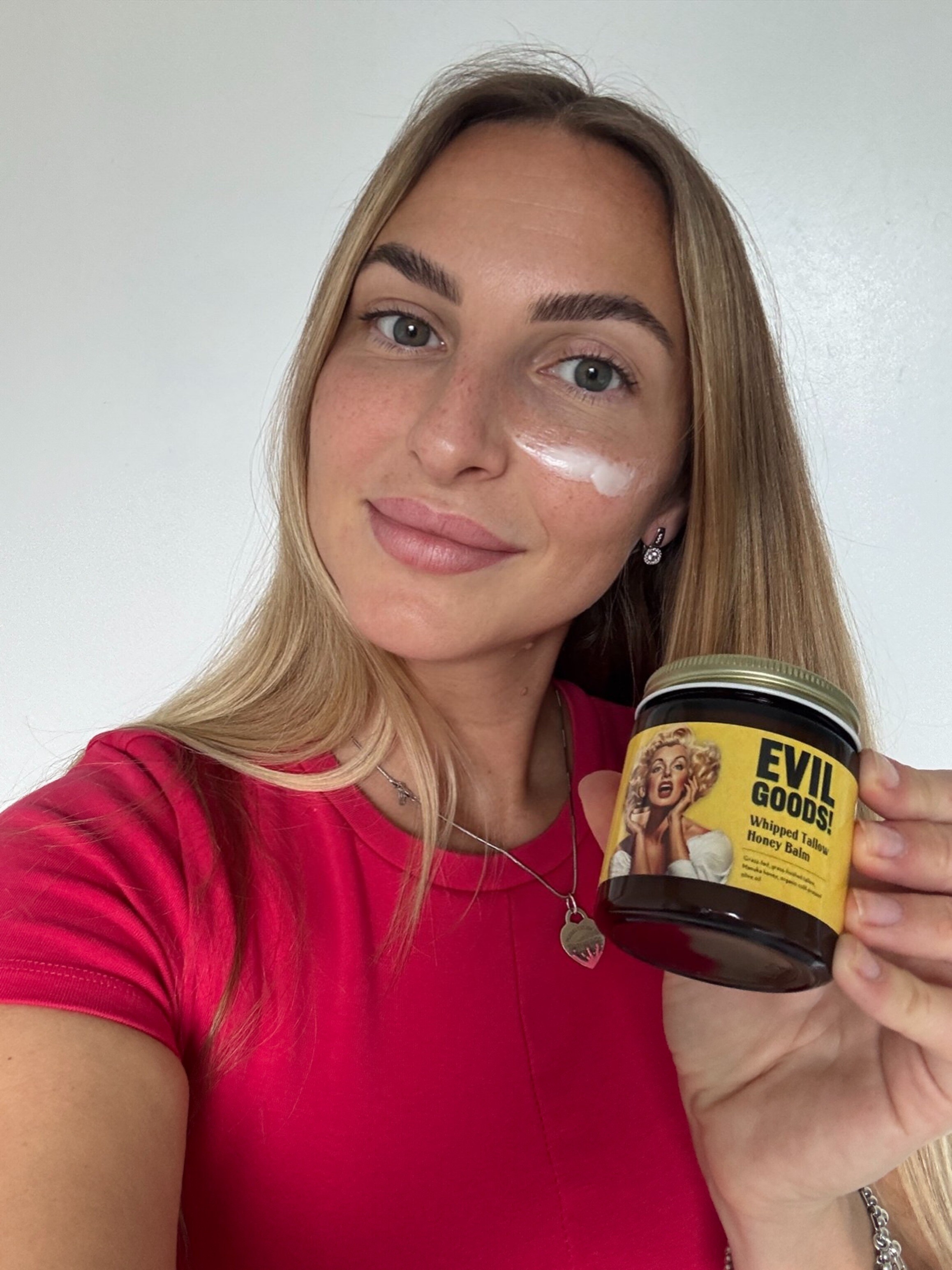 Person holding a jar labeled 'Evil Goods' with cream on their cheek.