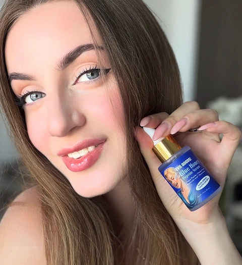 A smiling woman with long brown hair holds a small blue bottle of Evil Goods Blue Honey Peptide Serum.