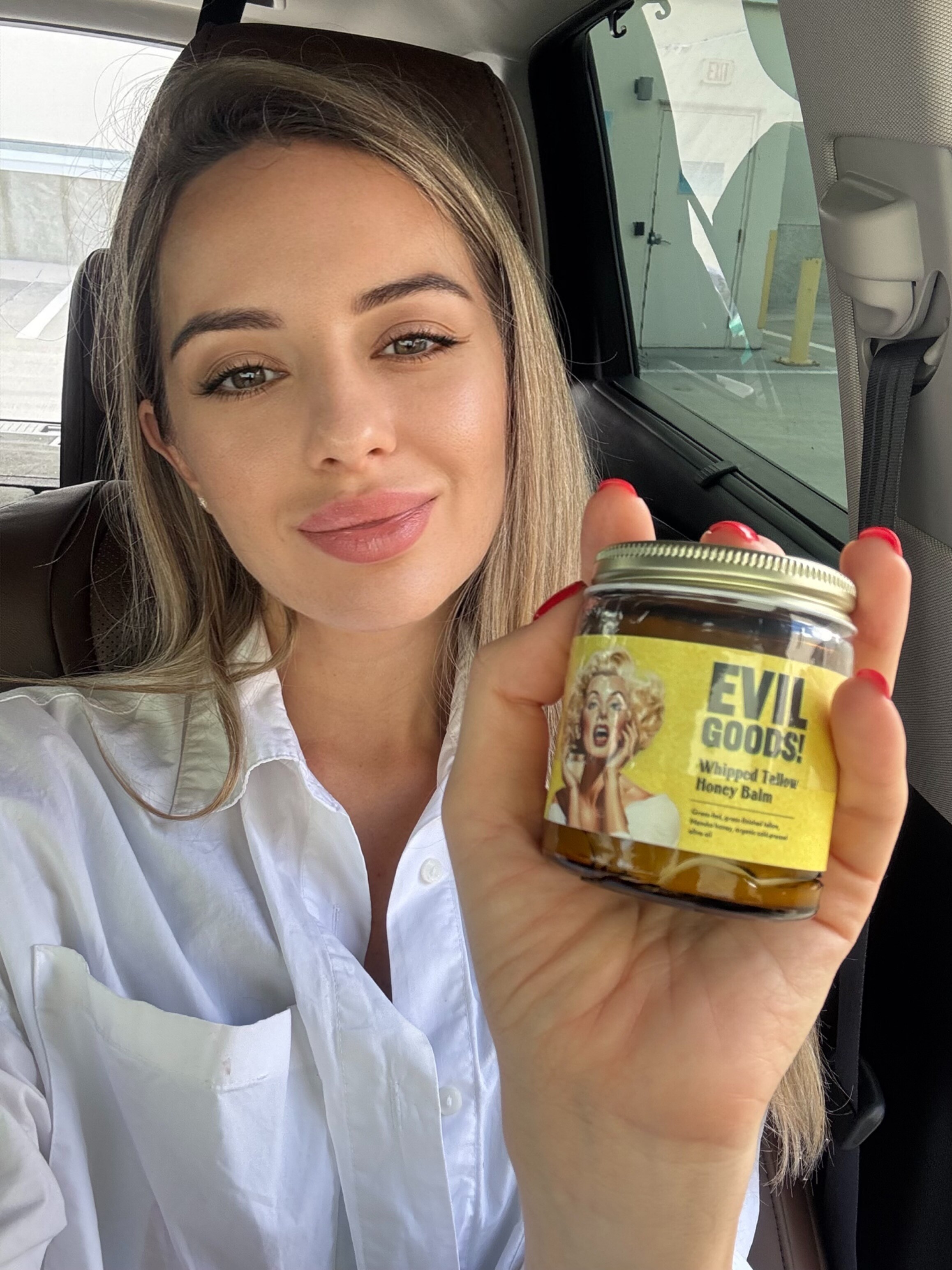 Person in a car holding a jar labeled 'Evil Goods Whipped Tallow Honey Balm'.