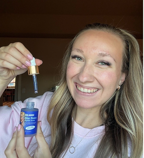 A smiling woman with blonde hair holds a small blue bottle of serum and its dropper over the opening.