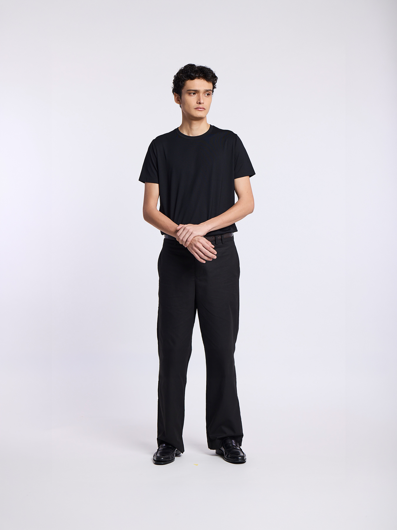 A man in a black t-shirt and black pants stands against a white background, looking to the side.