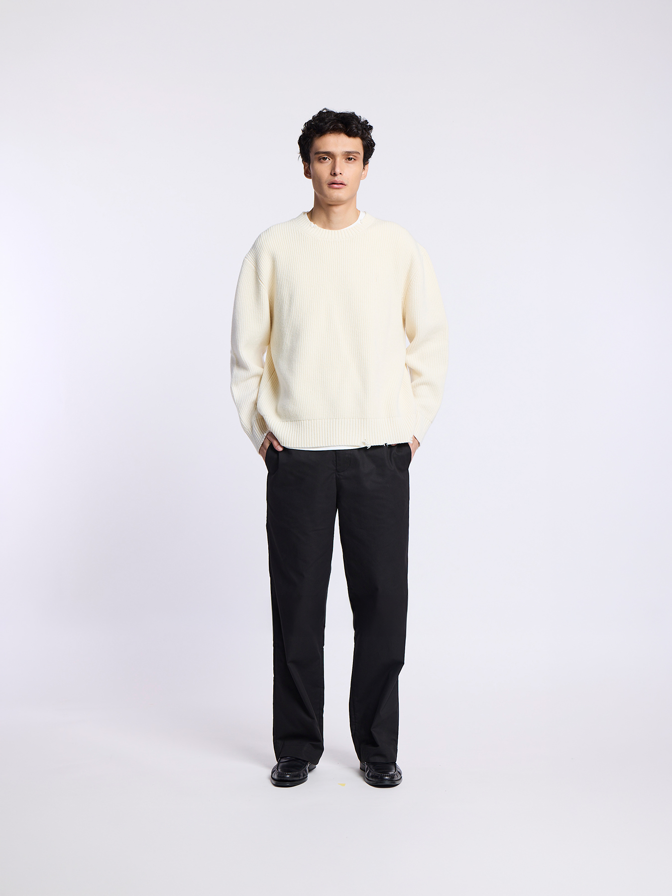 A man wearing a cream-colored ribbed sweater and black pants stands facing forward against a white background.