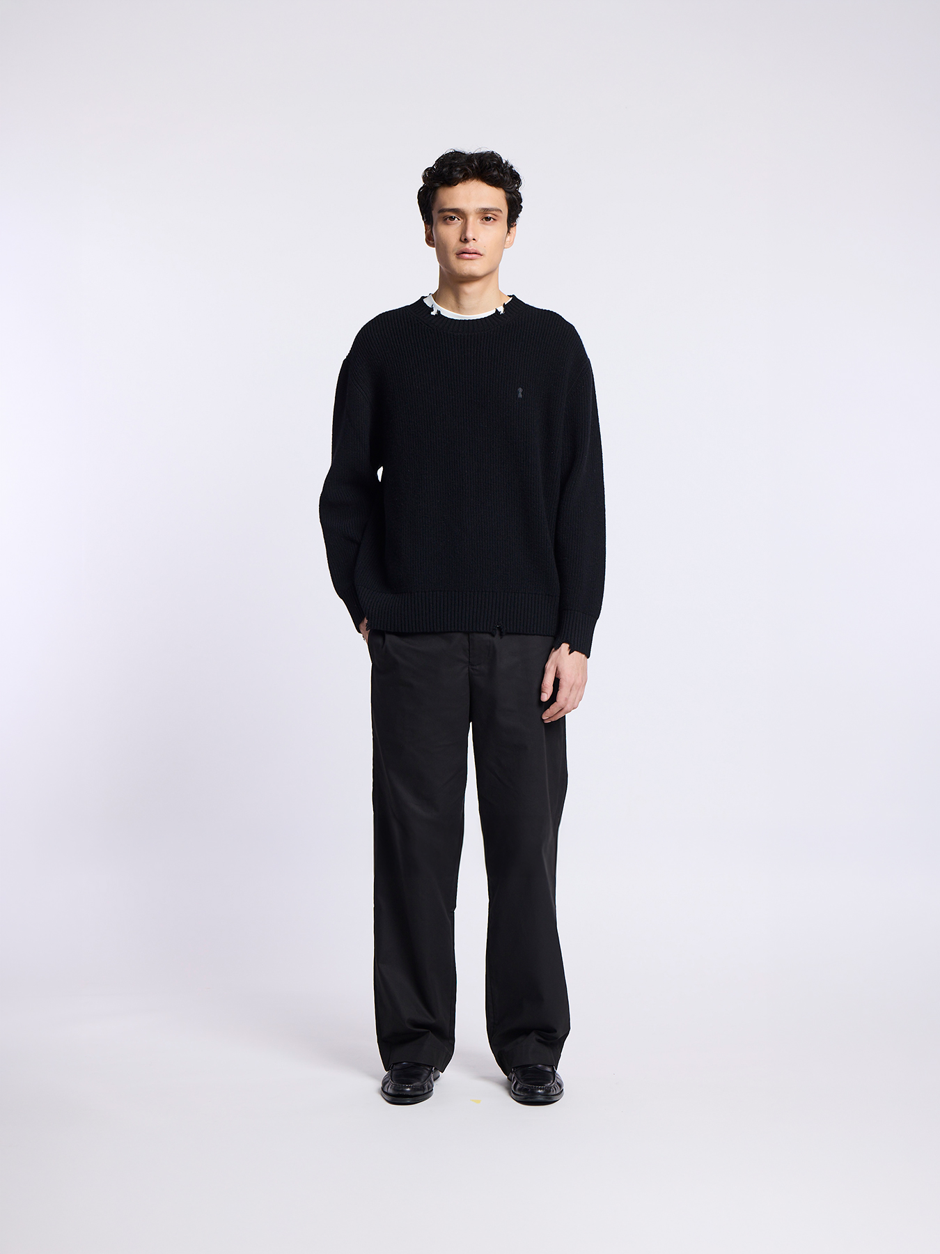 A man wearing a black sweater, black pants, and black shoes stands against a white background.