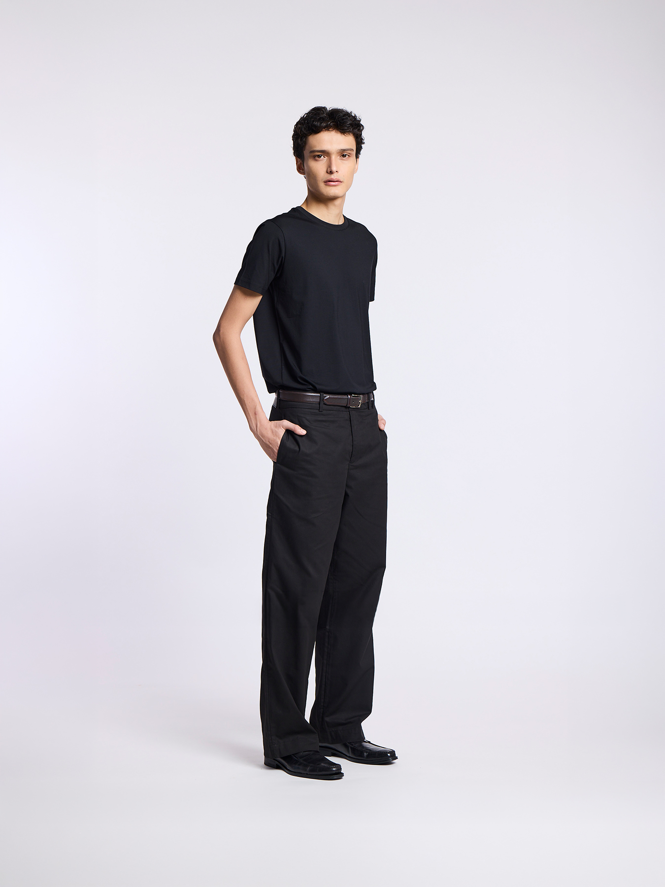 A man wearing a black t-shirt and black trousers poses against a plain white background.