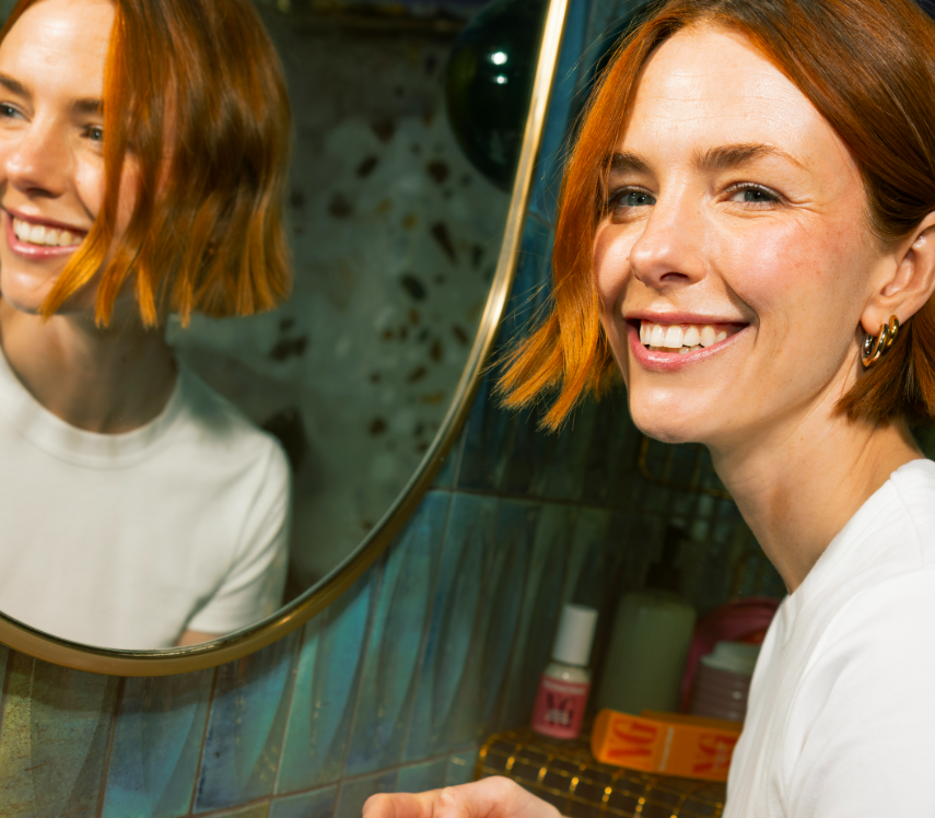 A smiling person with orange hair looking in a mirror.