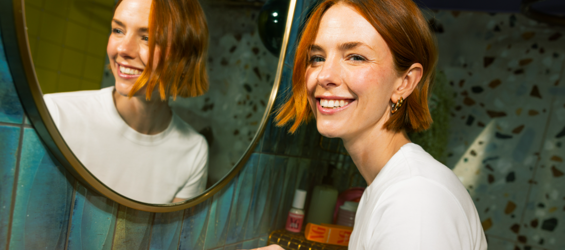 A person with short red hair smiling in front of a round mirror.