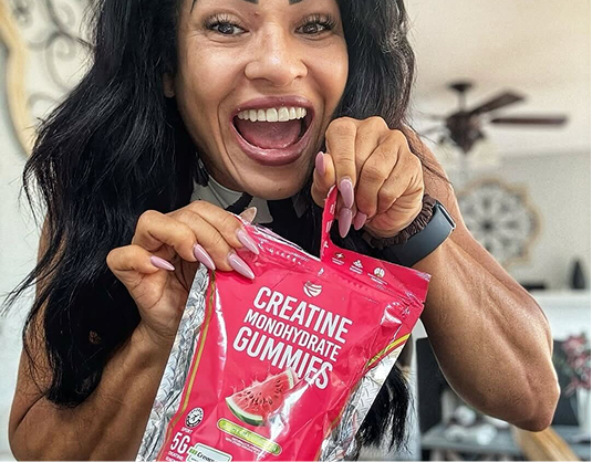 A muscular woman with long dark hair excitedly opens a red package of creatine monohydrate gummies.
