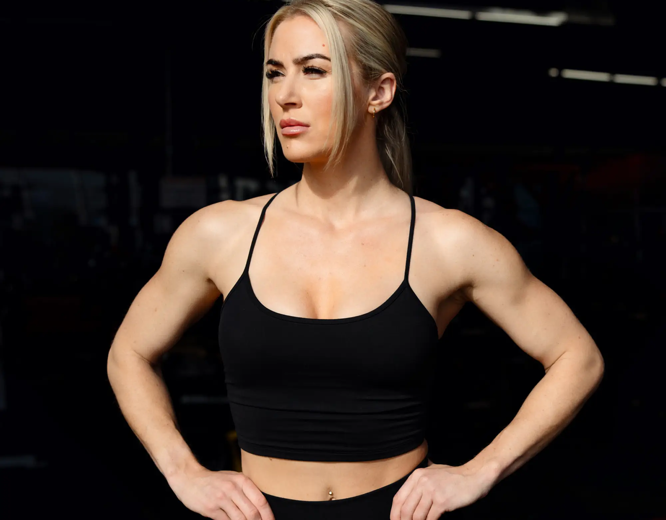 A fit, blonde woman in a black workout top with her hands on her hips, looking sideways.