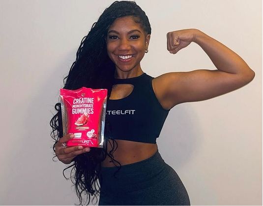 A smiling woman in workout clothes flexes her bicep while holding a package of creatine gummies.