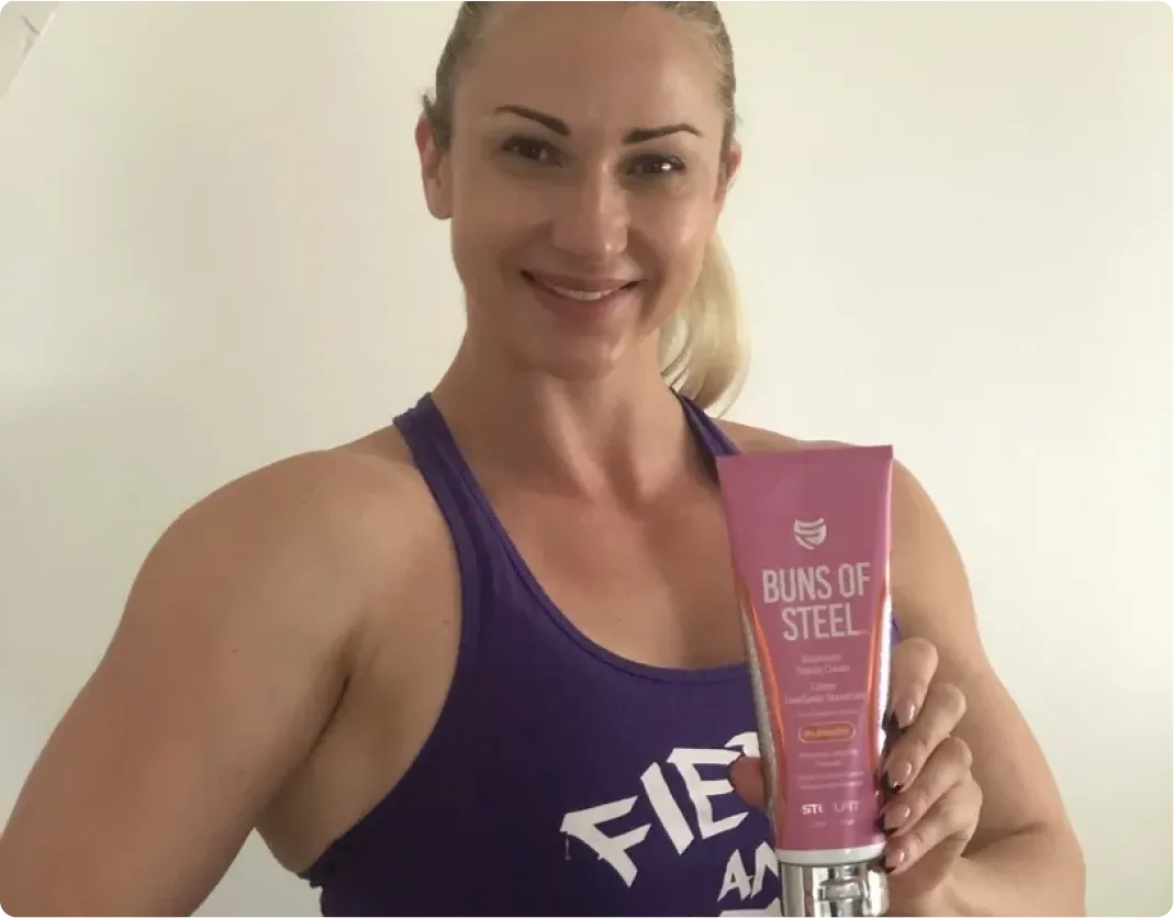 A woman in a purple tank top smiles while holding a pink tube of Buns of Steel lotion.