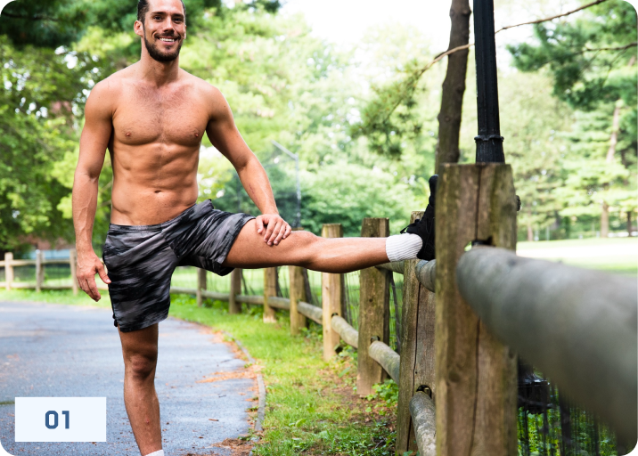 A shirtless, muscular man stretches his leg on a wooden fence in a park.