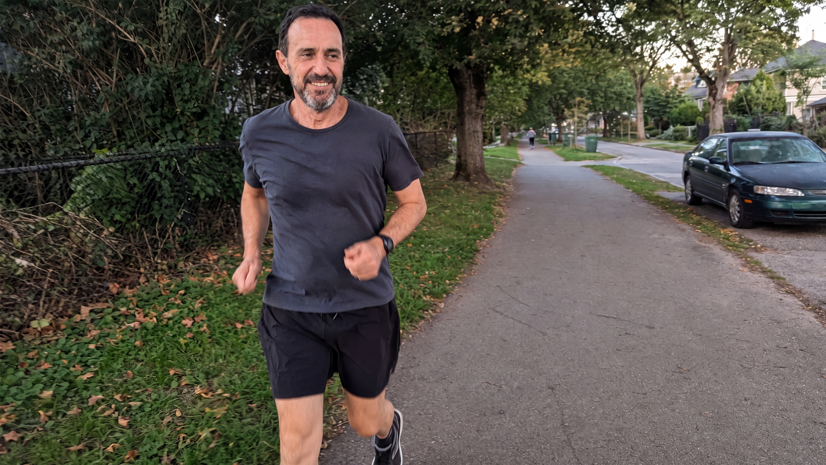 A smiling man with a beard jogging on a paved path next to a tree-lined street.