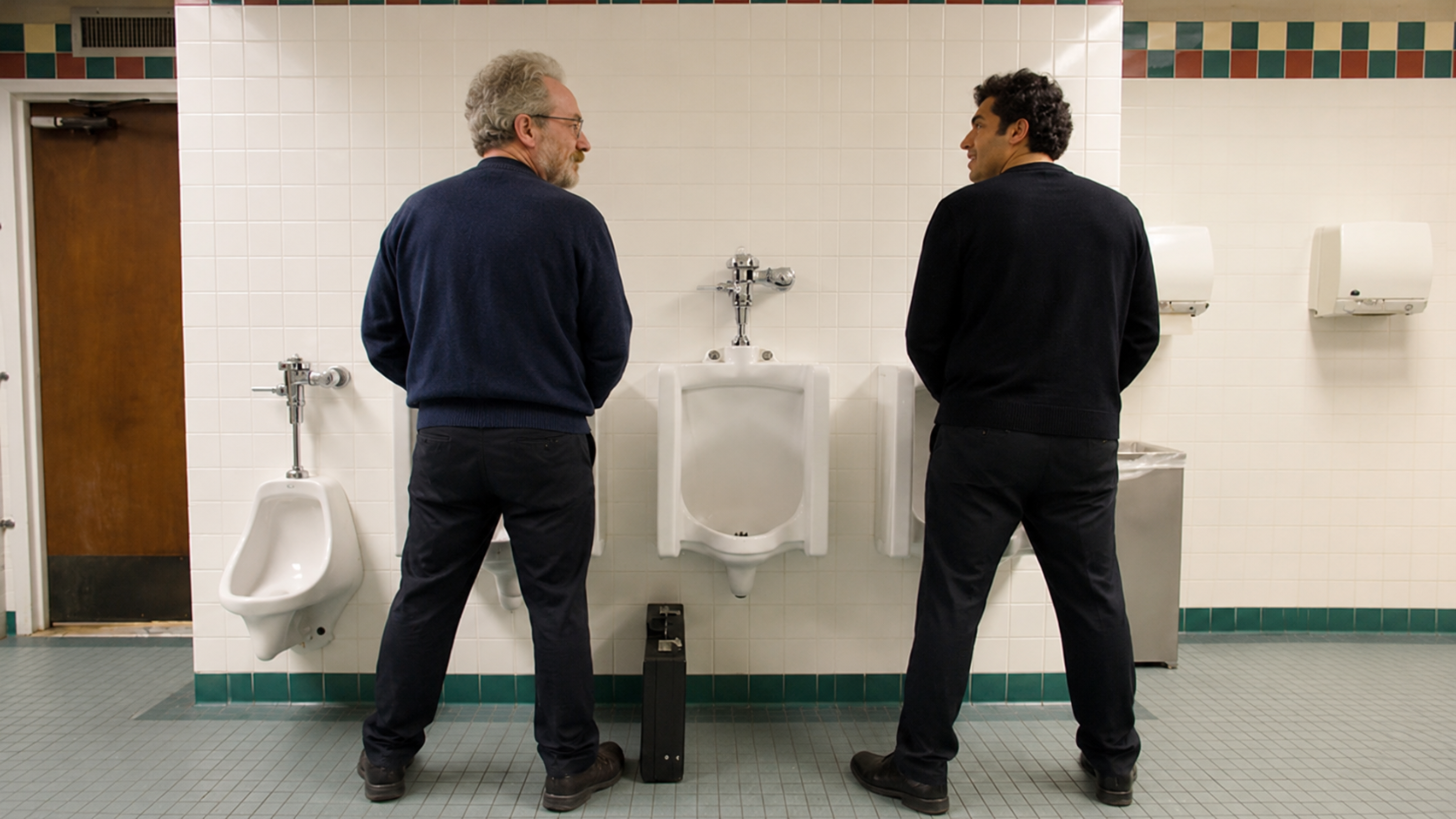 Two men stand at adjacent urinals in a public restroom, turned towards each other as if in conversation.