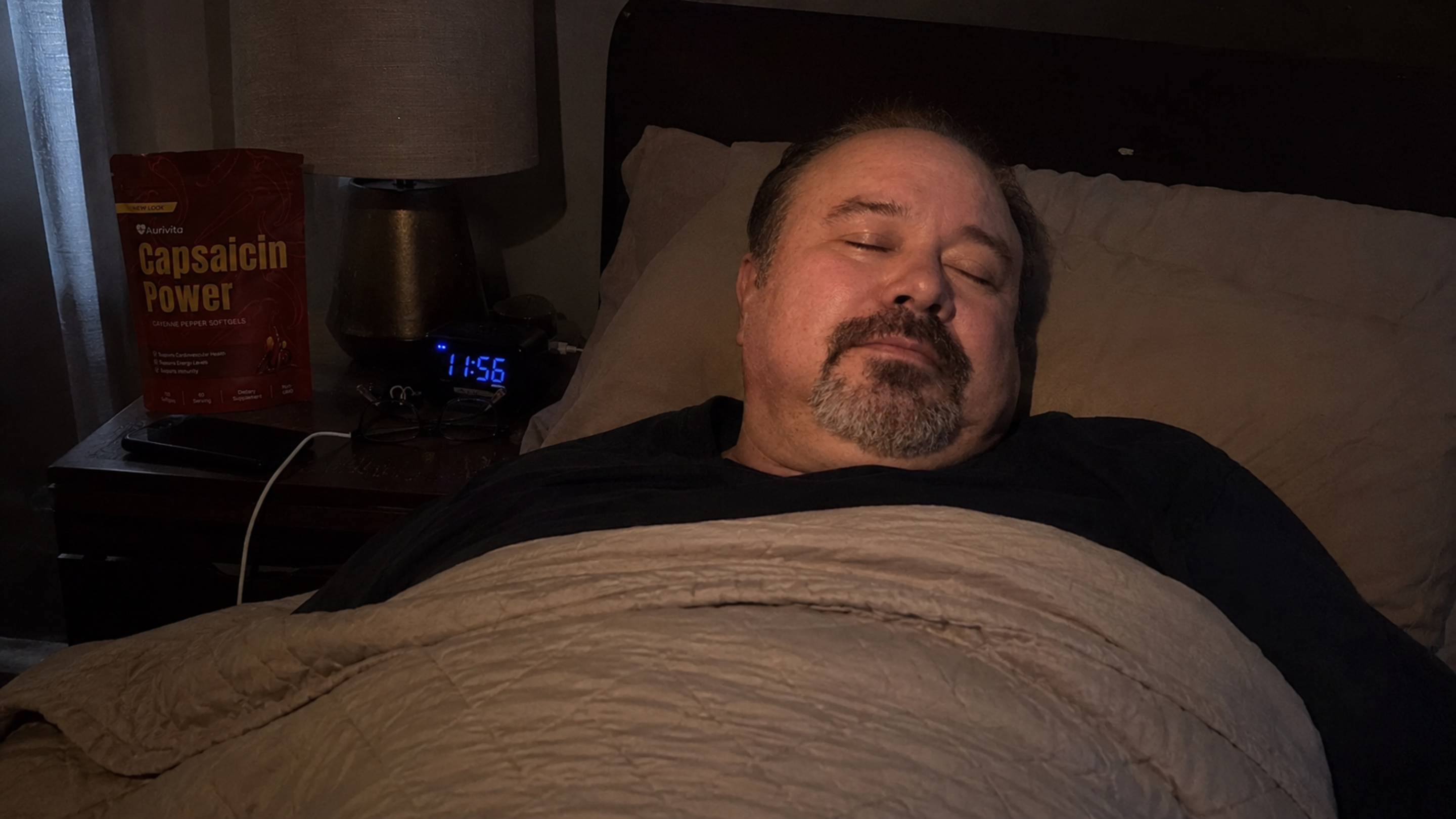 A man with a goatee sleeps in a dimly lit bedroom with a pouch of supplements on his nightstand.