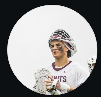 A young male lacrosse player in a helmet and jersey with eye black, holding his stick.