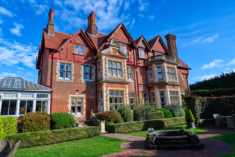 A large, red brick manor house with a manicured garden under a blue sky on a sunny day.