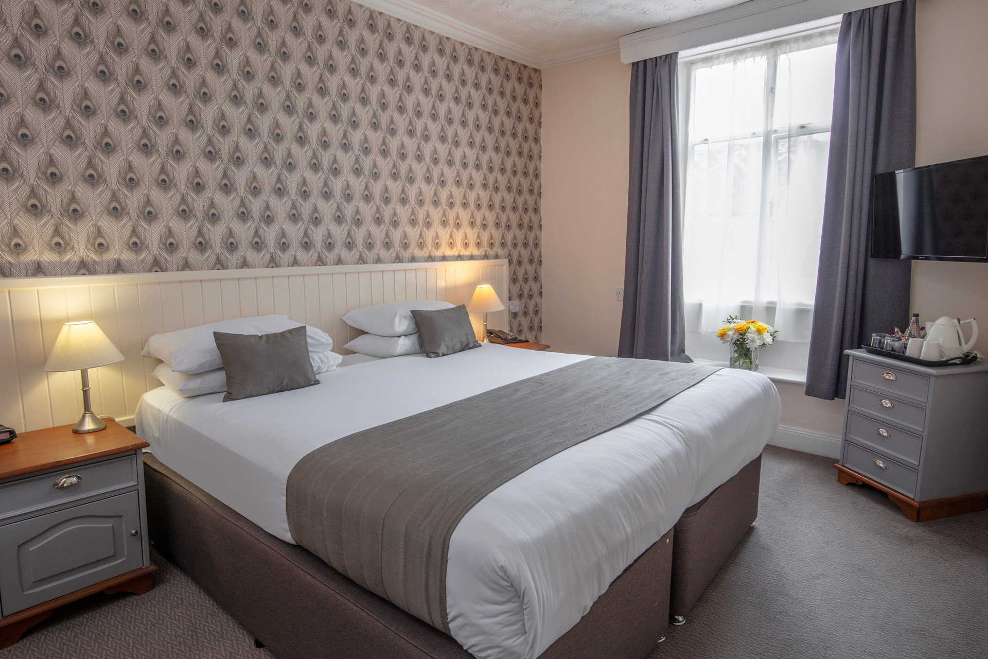 A hotel bedroom with a neatly made double bed, peacock feather patterned wallpaper, and a large window.