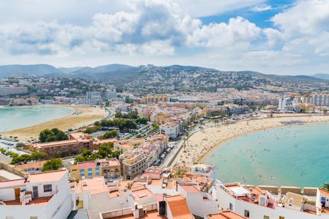 A high-angle view of a coastal city with two sandy beaches, turquoise water, and mountains in the background.