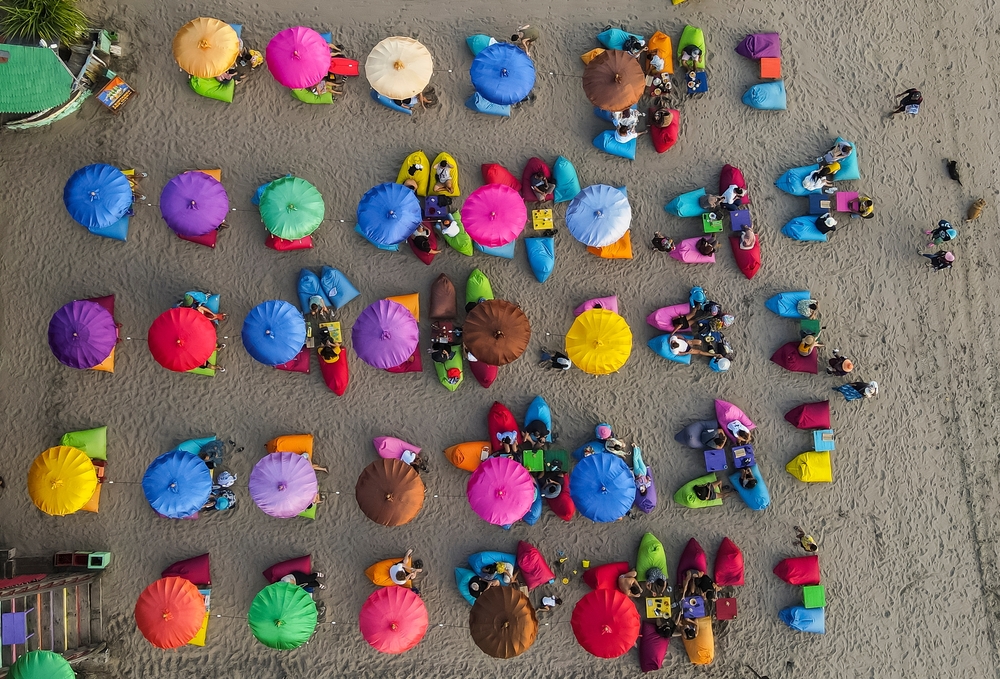 An aerial top-down view of a sandy beach with people sitting under many colorful umbrellas.