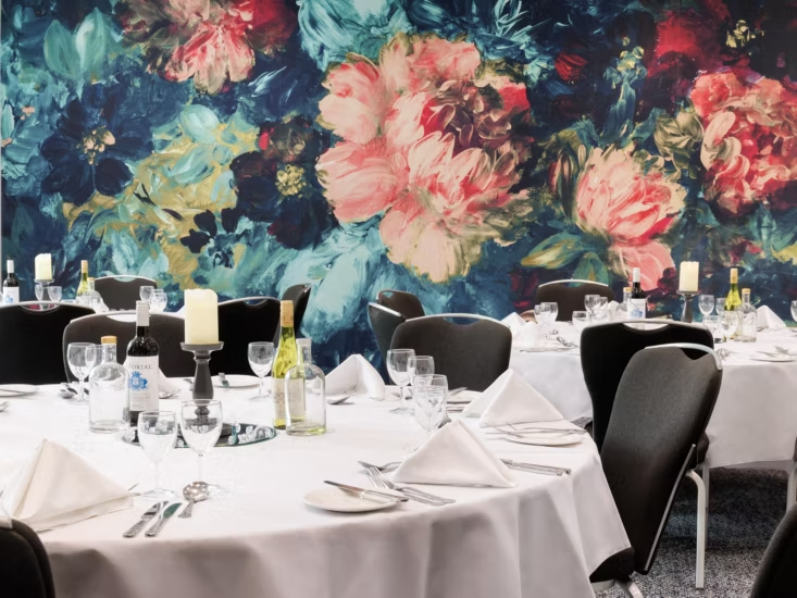Round dining tables set with white tablecloths and wine in front of a large, colorful floral mural.