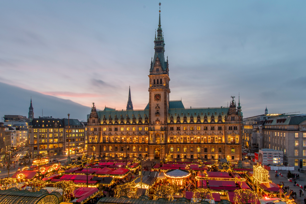 A brightly lit Christmas market in a city square in front of a large, historic building at dusk.