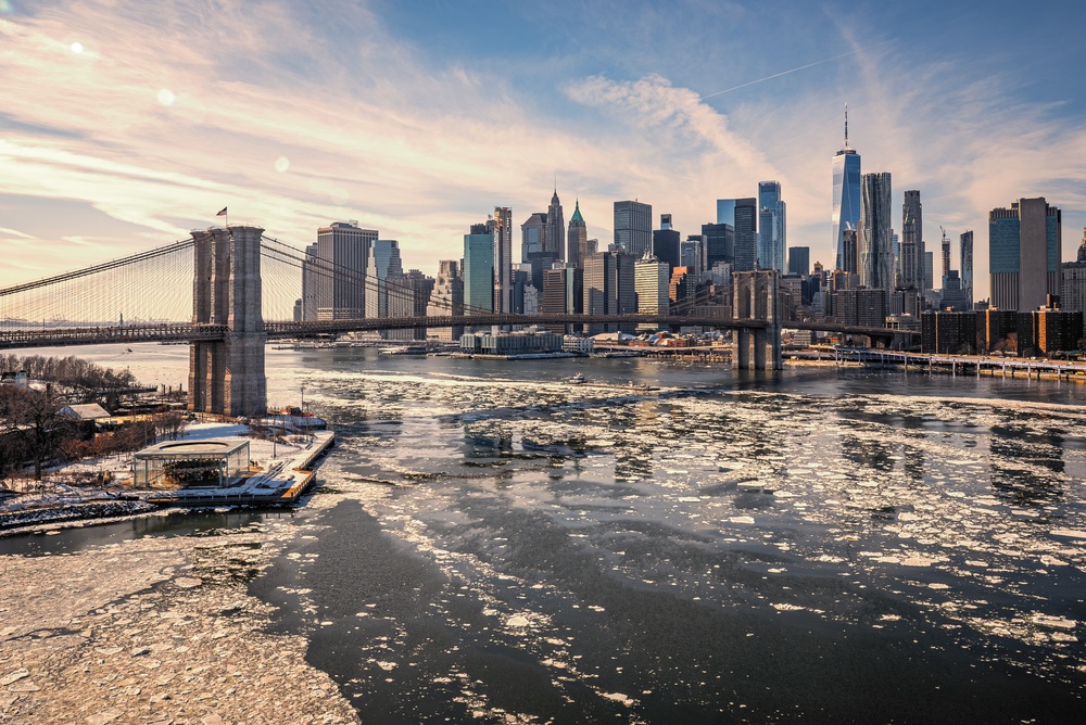 A winter view of the Brooklyn Bridge and the Manhattan skyline with ice floating on the river.