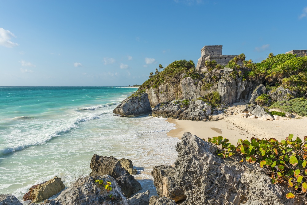 Stone ruins on a rocky cliff overlooking a tropical beach with turquoise water.