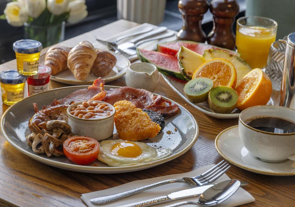 A full English breakfast spread on a wooden table with fresh fruit, croissants, coffee, and juice.