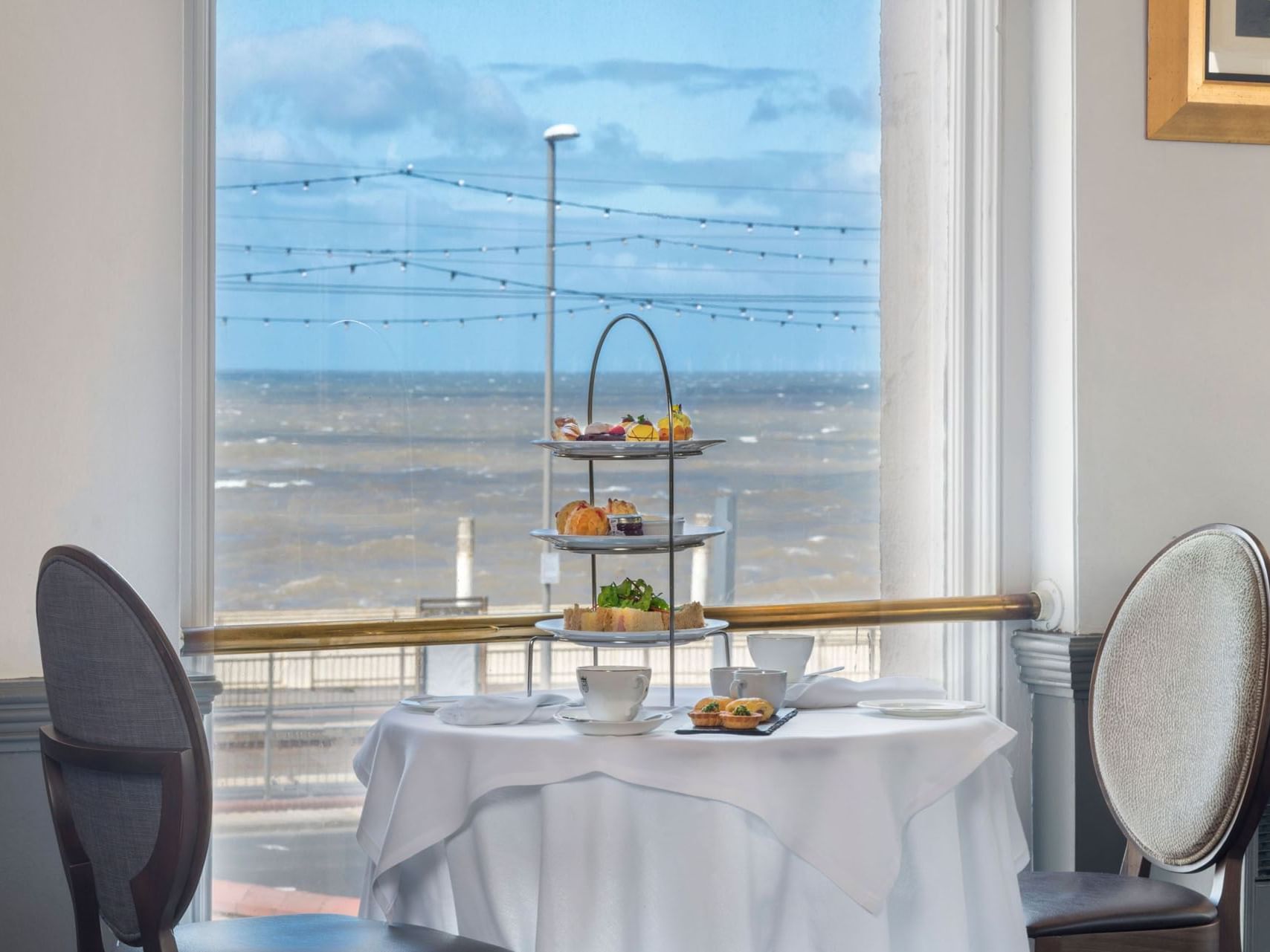 A table set for afternoon tea with a three-tiered stand by a window overlooking the sea.