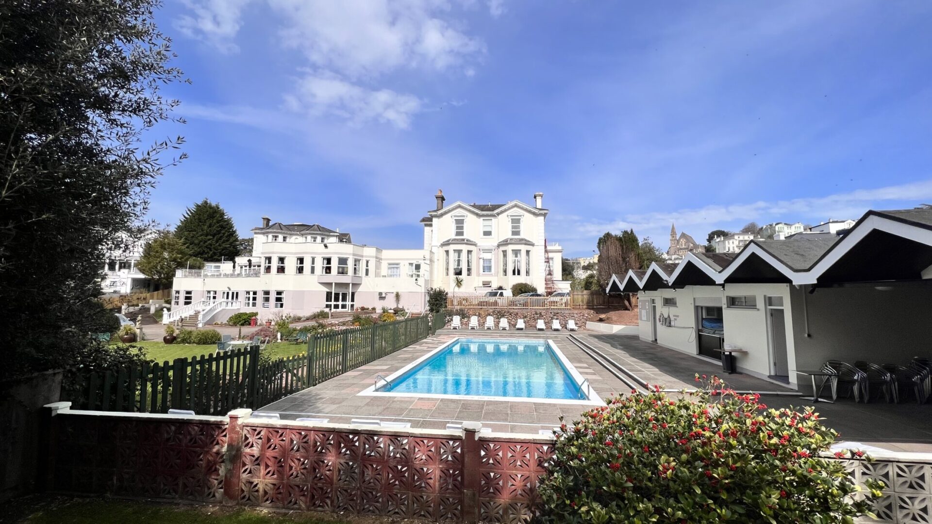 A large white hotel with an outdoor swimming pool and patio on a sunny day.