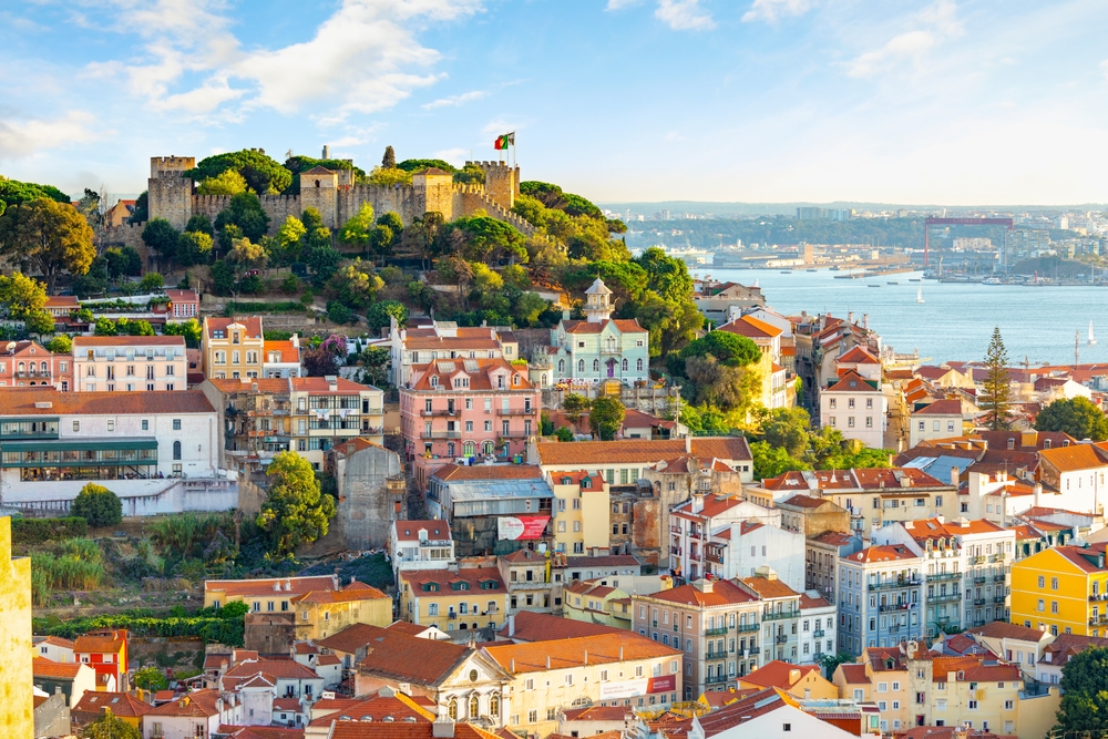 A sprawling city with colorful buildings and a stone castle on a hill overlooking the sea.
