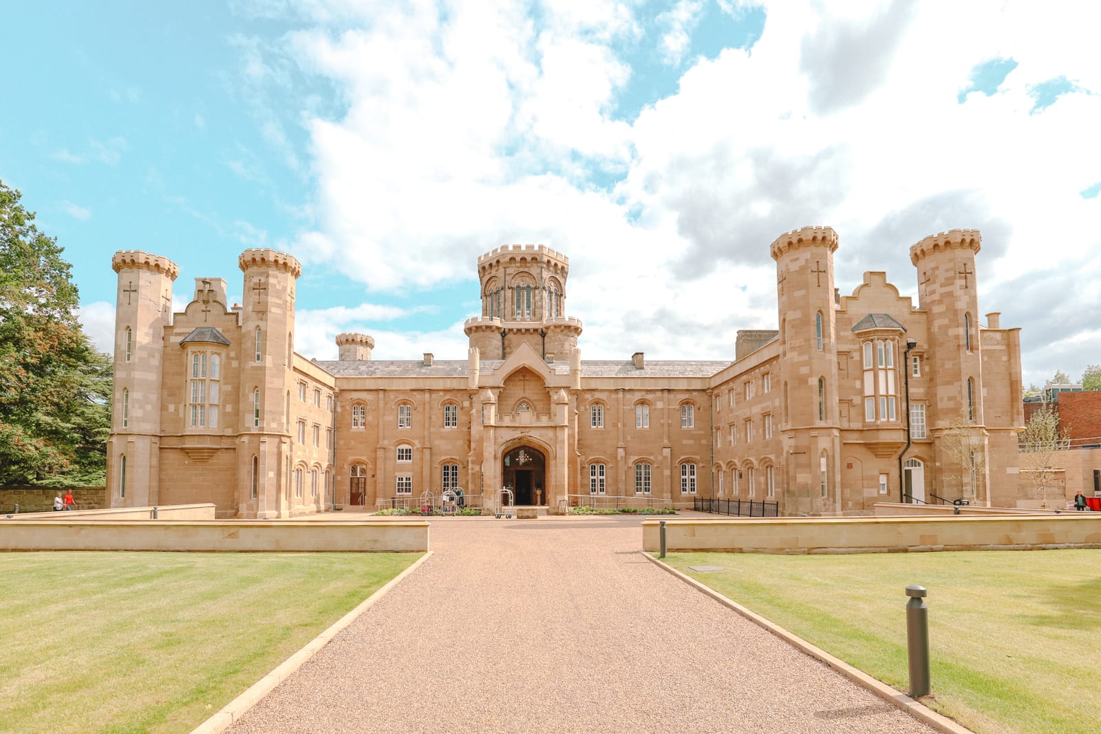 A large, stone castle-like building with a central gravel path leading to the entrance under a cloudy sky.