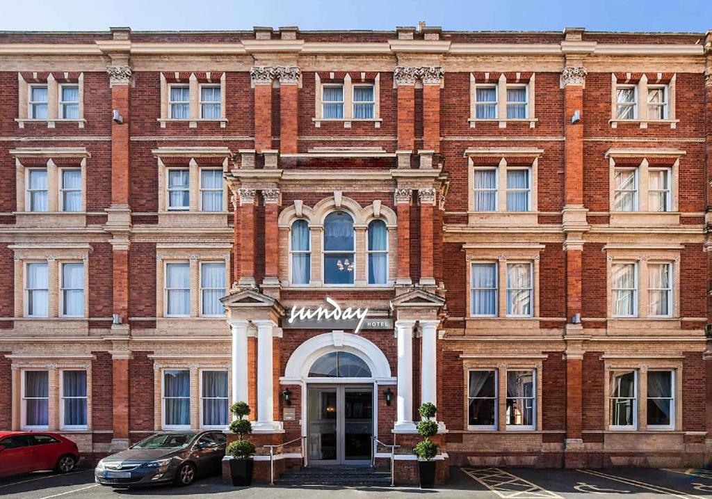 The ornate red brick facade of the Sunday Hotel on a clear day, with cars parked in front.