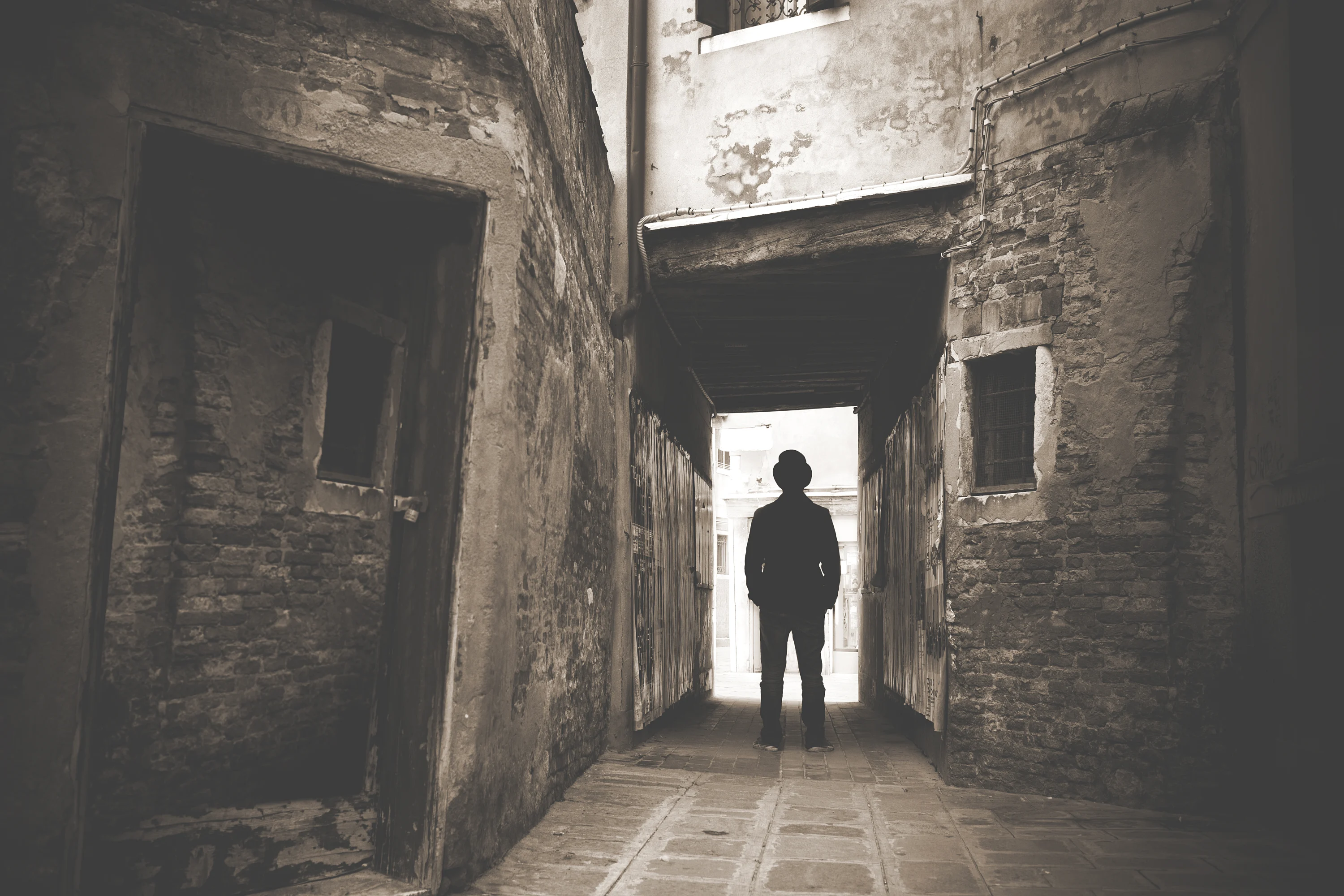 A black and white photo of a person's silhouette in a hat standing in a narrow alleyway.