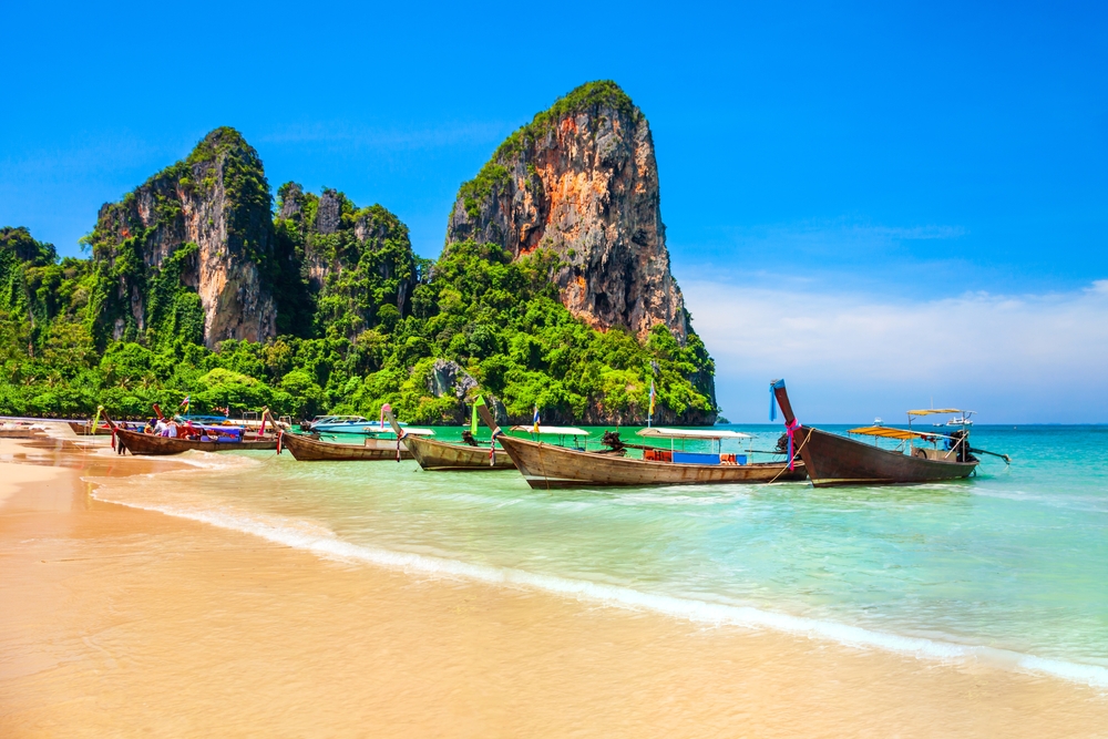 Traditional long-tail boats line a tropical beach with towering green limestone cliffs in the background.