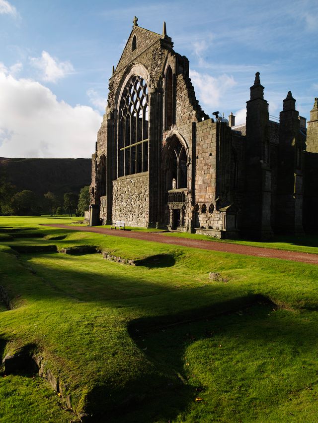 The stone ruins of a Gothic abbey stand in a green field with long shadows from the sun.