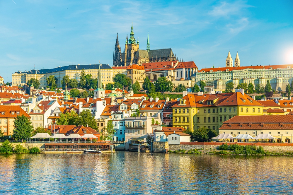 A sunlit view of a historic European city with a castle on a hill, seen from across a river.
