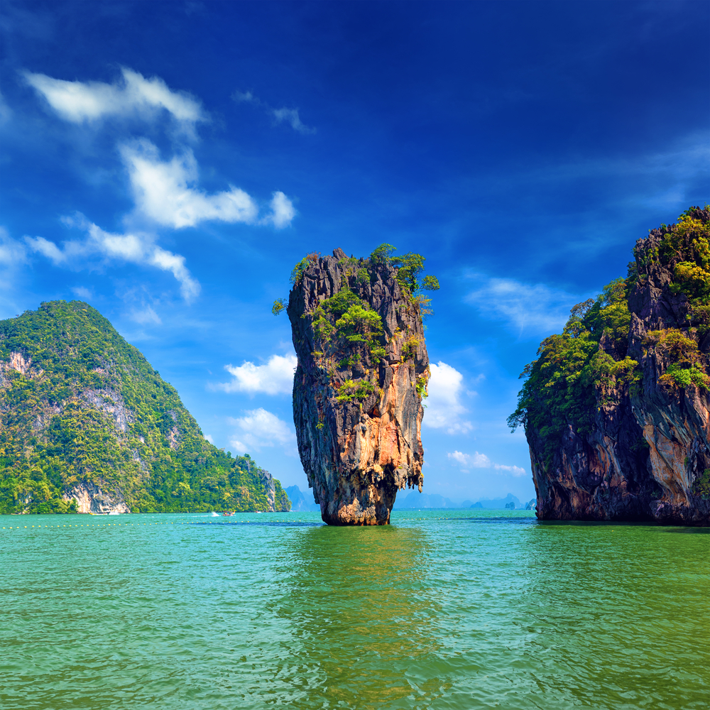 A tall, tree-topped rock formation stands in green water with other islands under a blue sky.