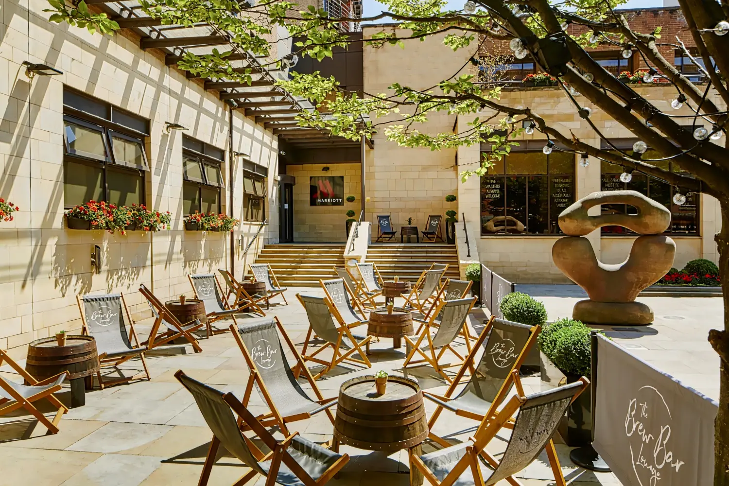 A sunny hotel courtyard patio with deck chairs arranged around barrel tables, with stairs leading up to an entrance.
