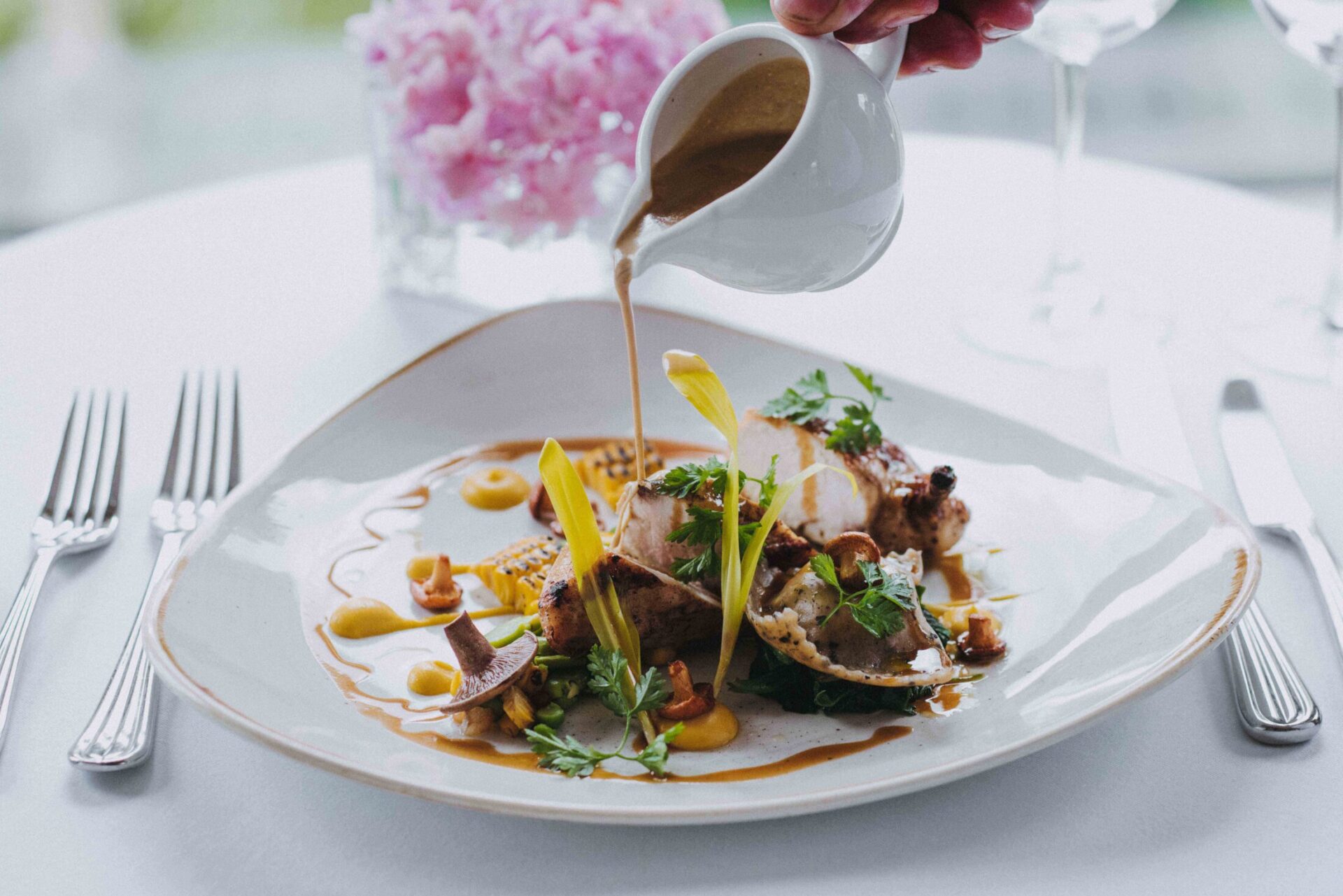 A hand pours sauce from a small pitcher onto a gourmet dish of roasted meat and vegetables.