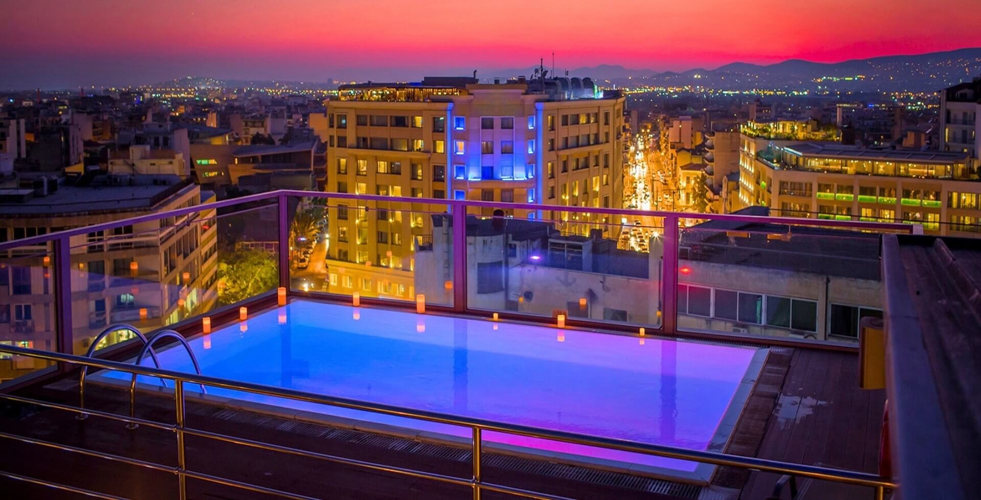 A rooftop swimming pool surrounded by candles, overlooking a city skyline at dusk with a vibrant pink sky.