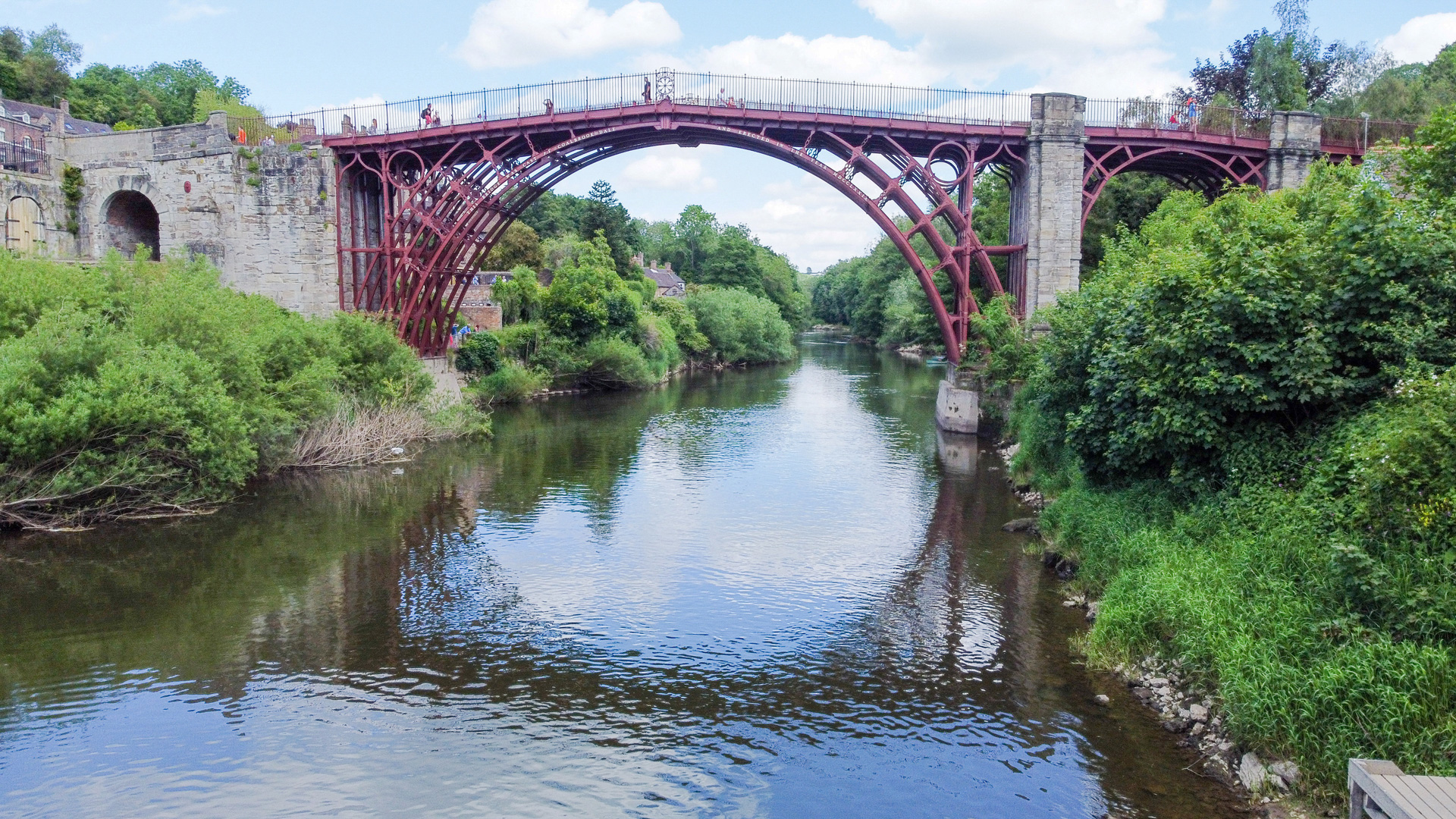 A wide, red, cast-iron arched bridge spans a river surrounded by lush green trees under a blue sky.