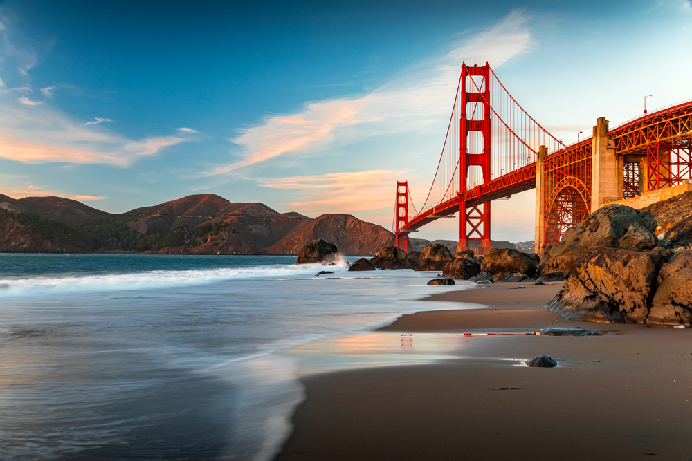 The Golden Gate Bridge viewed from a rocky beach at sunset, with waves washing ashore.