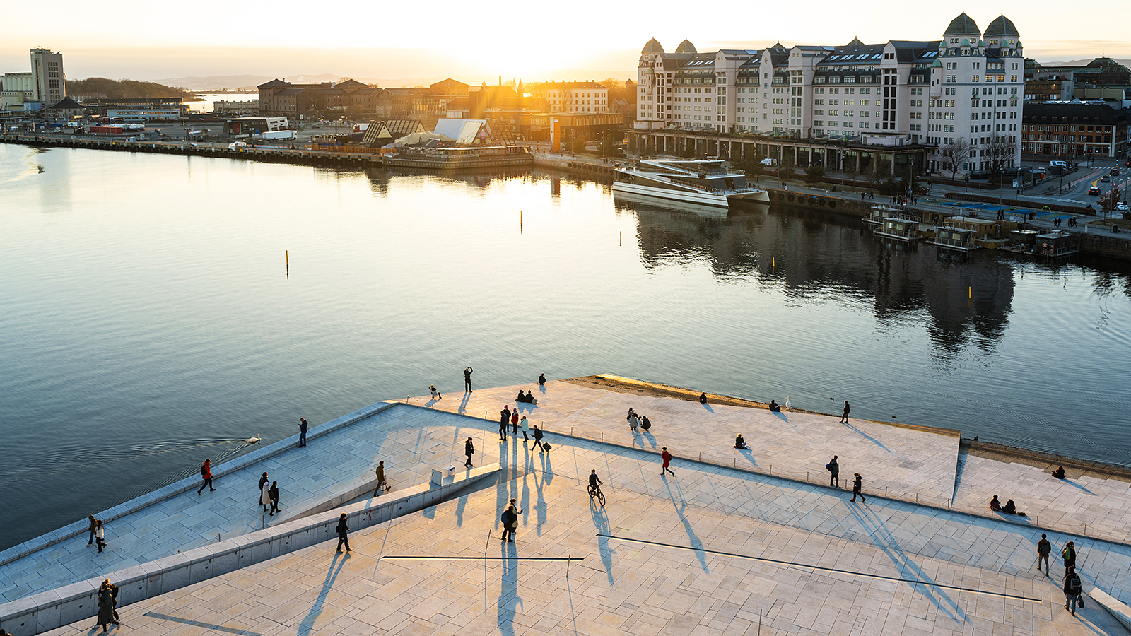 An elevated view of a city harbor at sunset, with people walking on a large stone plaza.