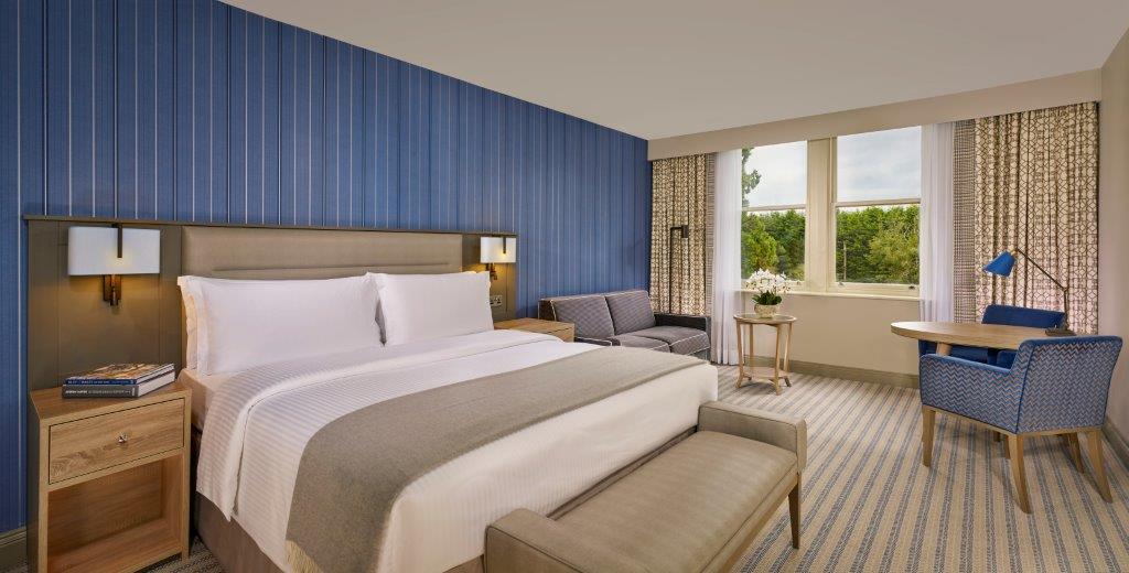 A tidy hotel room with a bed, blue striped wallpaper, a small sofa, and a desk by the window.