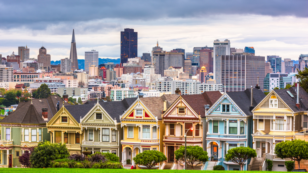 A row of colorful Victorian houses with the San Francisco city skyline in the background under a cloudy sky.