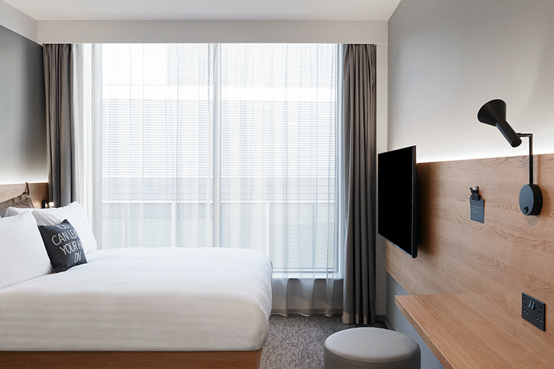 A modern hotel room with a white bed, a large window with sheer curtains, and a wall-mounted TV.